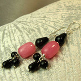 A pair of handmade dangle earrings featuring large pink teardrop beads, smaller black teardrop beads, and small black round beads on silver-toned French hook wires, resting on a textured light gray fabric surface.